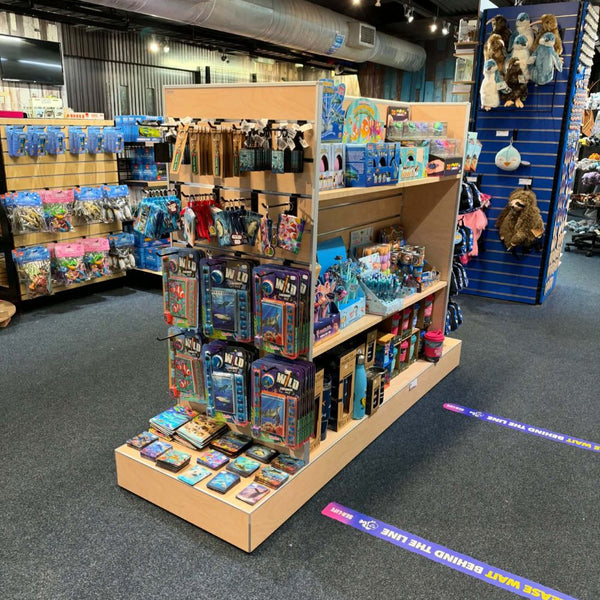A retail display slatwall stand in natural wood filled with assorted toys, books, and packaged merchandise inside a store. The stand has multiple shelves and hanging racks, with products like action figures, boxed items, and small accessories visible.