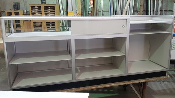 A white retail counter unit with a large glass display case and open shelving in a factory setting - back view