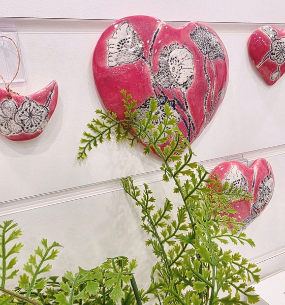 Pink heart-shaped ceramic decorations with silver floral patterns on a white surface with green leaves hung by a keyhole clip