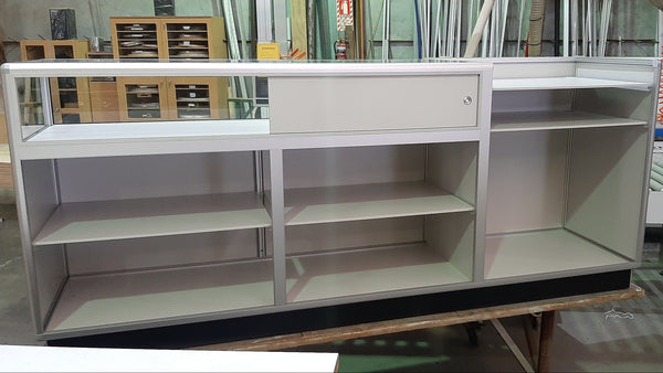 A white retail counter unit with a large glass display case and open shelving in a factory setting - back view
