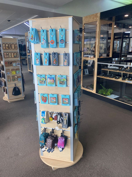 A freestanding slatwall retail display stand inside a store, holding multiple blue and multicolor zip pouches, small packaged items, and a few bags. The stand has a light wood top and base with a white frame, and the surrounding area includes carpeted floor, shelving, and other display fixtures.