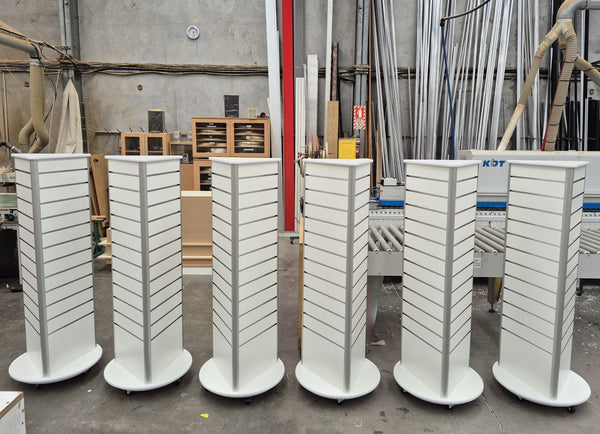 Six white slatwall 3 sided retail display stands arranged in a row inside a workshop setting with industrial equipment and materials visible in the background.