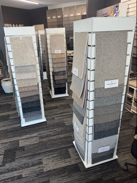 Three carpet sample slatwall display towers showing various textured floor options in beige, gray, and brown tones, set on a patterned gray carpet inside a showroom.