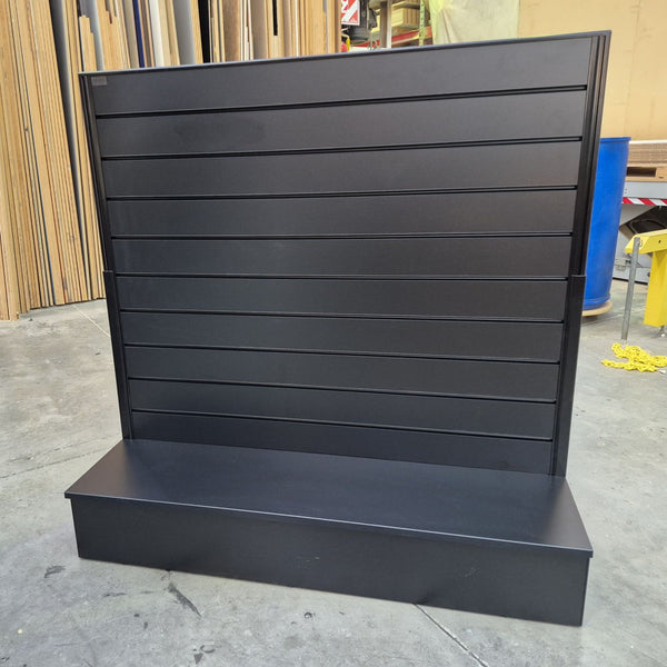 A black slatwall free standing light weight display stand with a base shelf, photographed in the Plankwall warehouse with concrete floors and wood panels in the background.